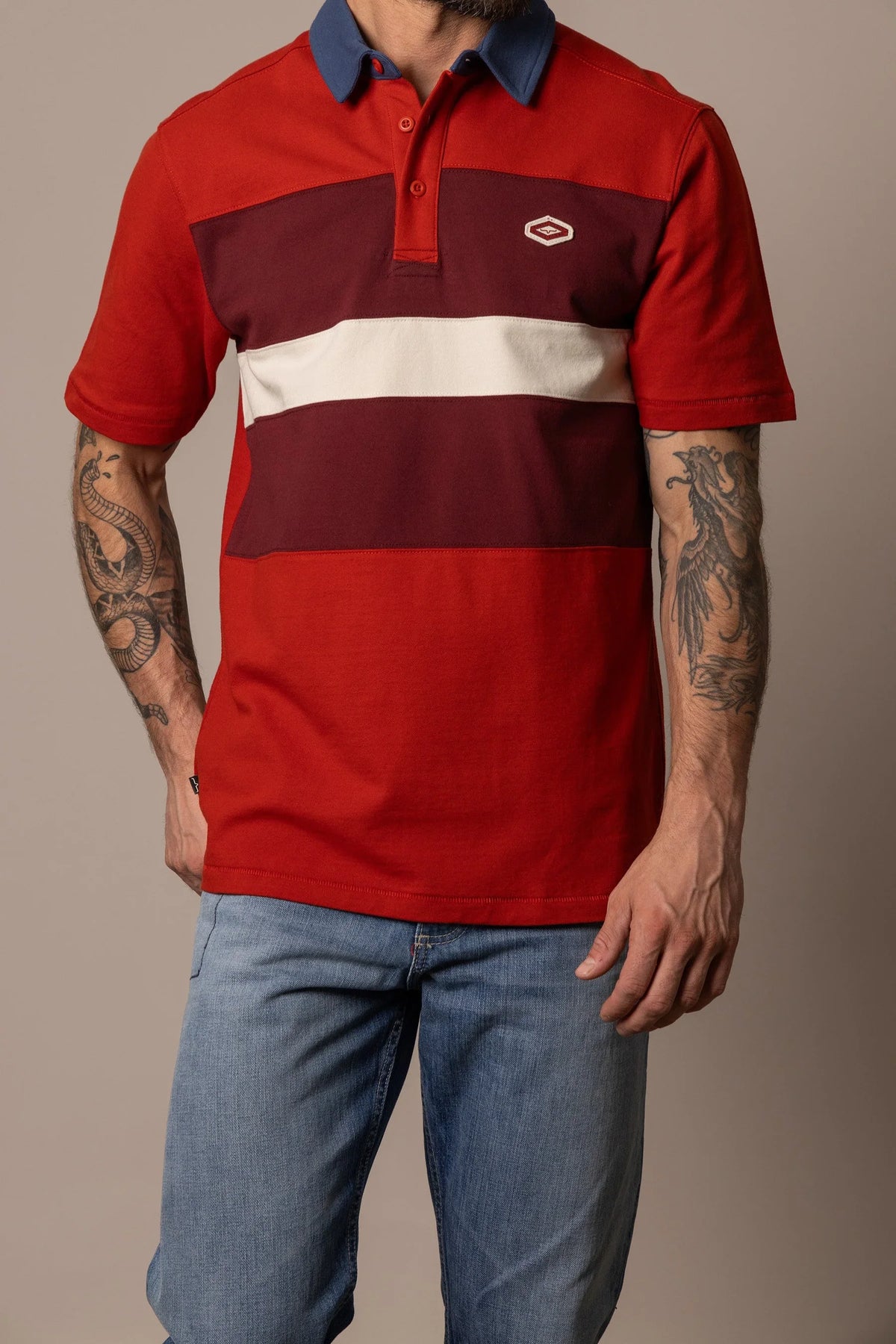 Kimes Ranch 1874 Polo in Brick Red with a navy collar and horizontal stripes in burgundy and cream and a logo on the chest