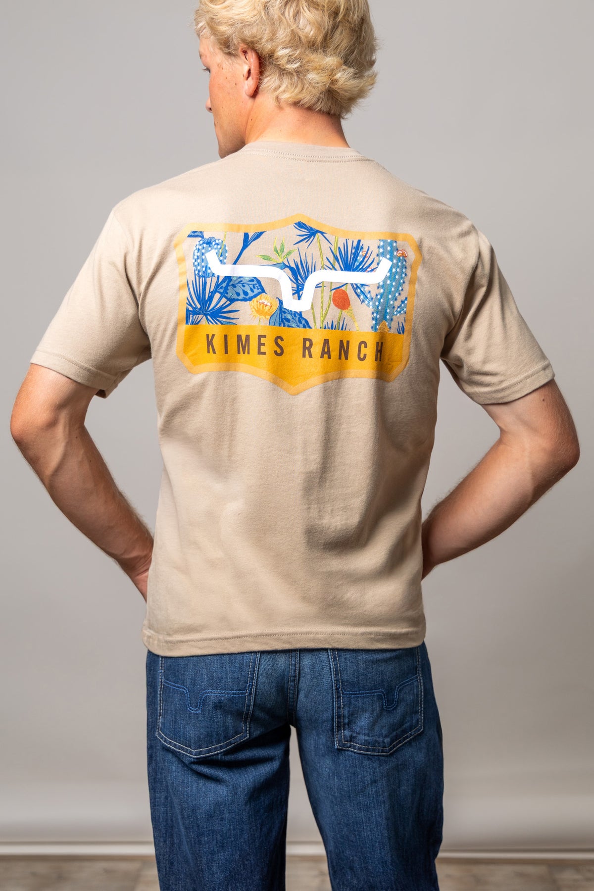 Kimes Ranch Dillard Floral shirt in Tan, back view, with a back graphic of blue tropical foliage and the Kimes Ranch logo