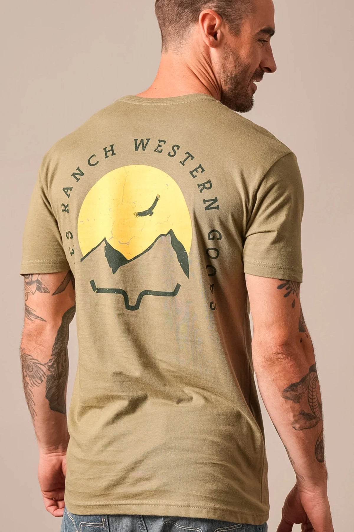 Kimes Ranch Flying High shirt in Light Olive, back view, with a back graphic of mountains and a bird under a yellow sun