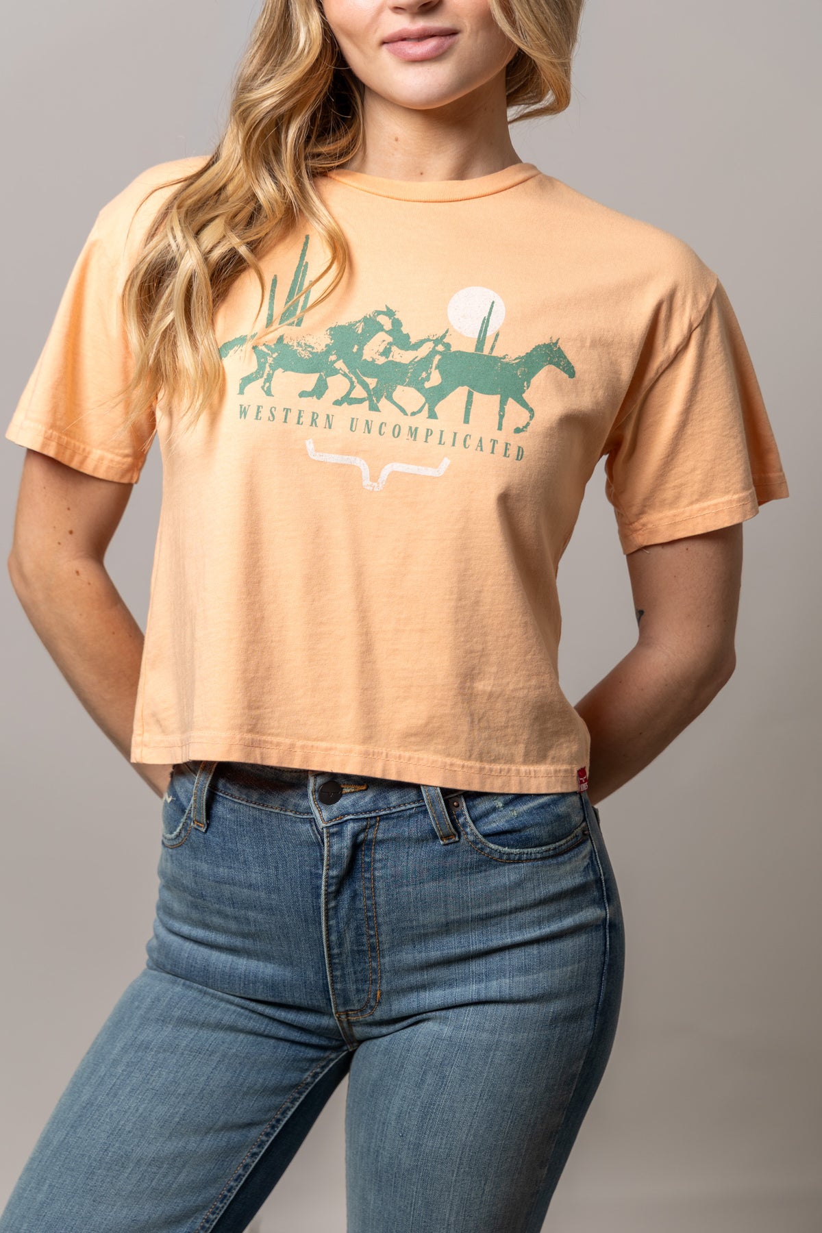 Kimes Ranch Plains Stampede shirt in Peach with a graphic of cowboys on horses and the text Western Uncomplicated
