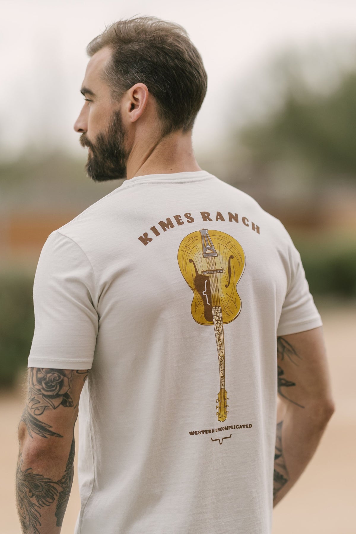 Kimes Ranch Lucky B Tuned shirt in Natural, back view, with a back graphic of a guitar and Western Uncomplicated text