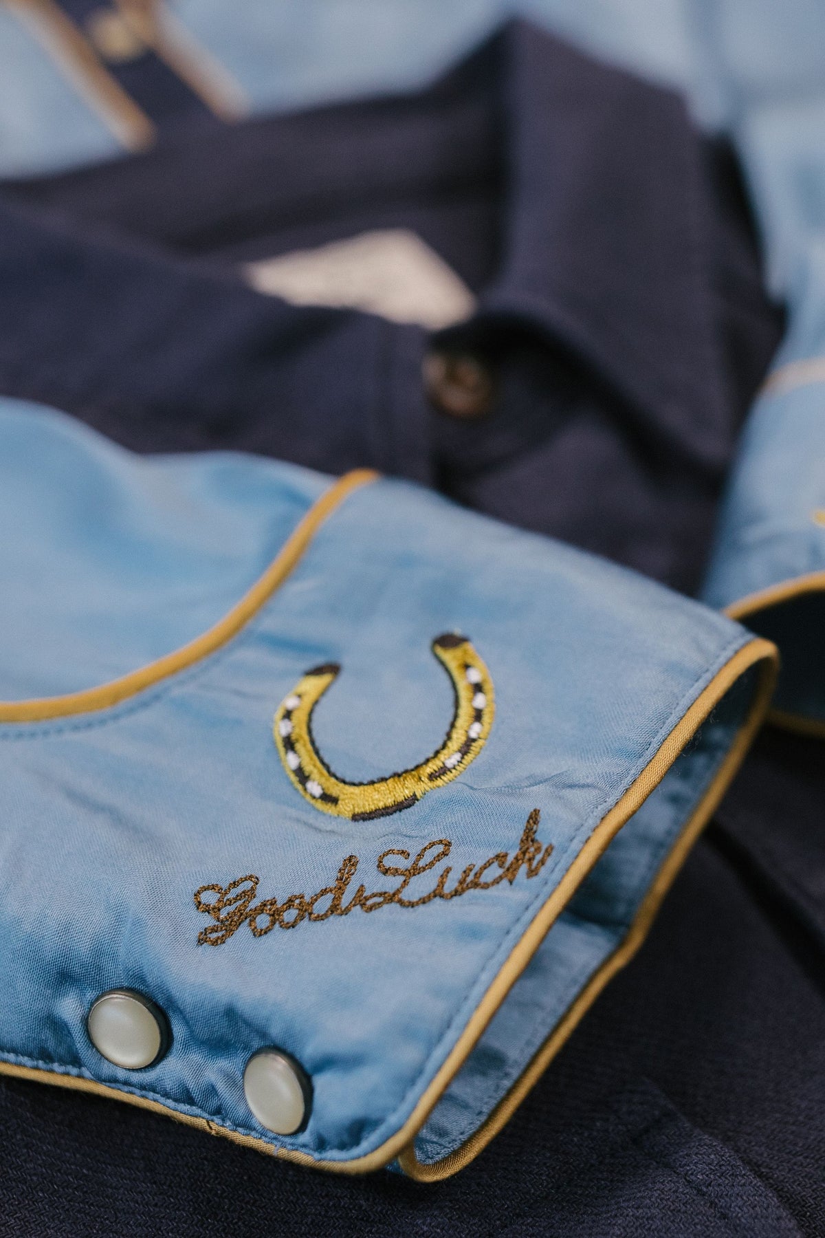 Kimes Ranch Merada top in Mid Blue, close-up, with embroidered horseshoe and Good Luck text on the cuff