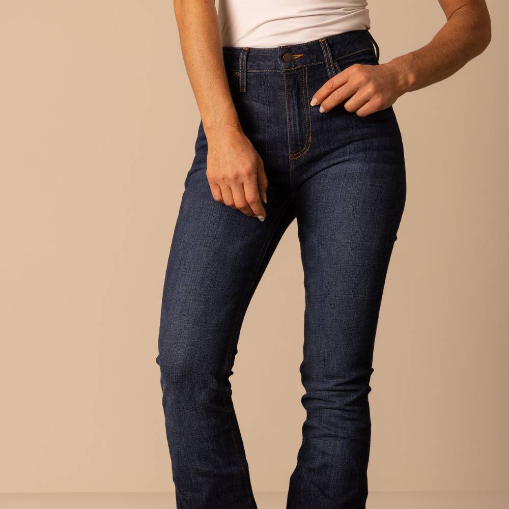 Woman wearing Kimes Ranch high-rise jeans in a dark indigo wash, styled with a white top. The jeans feature a flattering, slim fit through the hips and thighs with classic Western stitching