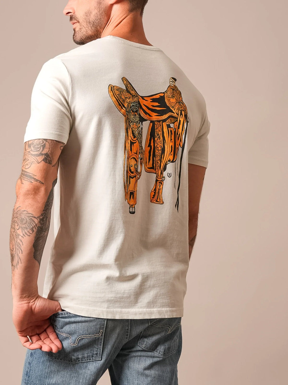 Kimes Ranch Lucky B Saddle short sleeve shirt in Natural, back view, with a large orange and black saddle graphic on the back