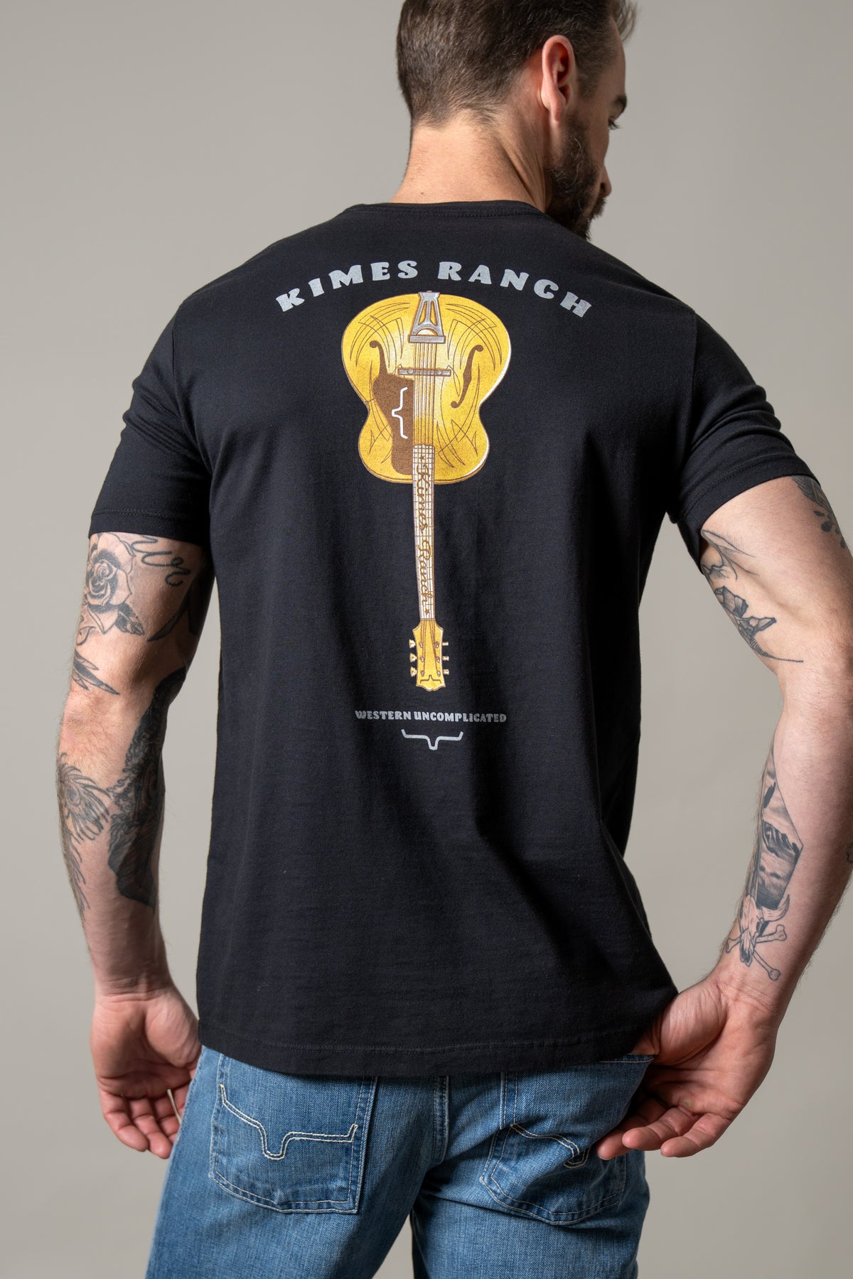 Kimes Ranch Lucky B Tuned shirt in Black, back view, with a gold acoustic guitar graphic and Western Uncomplicated text