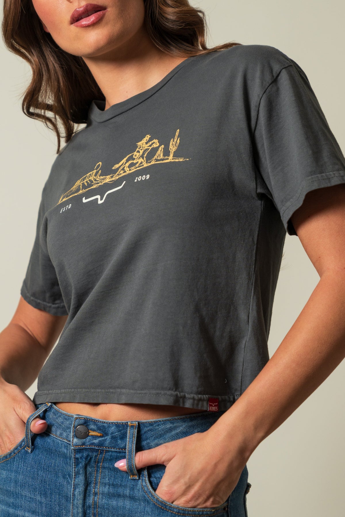 Kimes Ranch Go West cropped tee in Pepper, front view showing short sleeves and a gold graphic with riders, cacti, and text